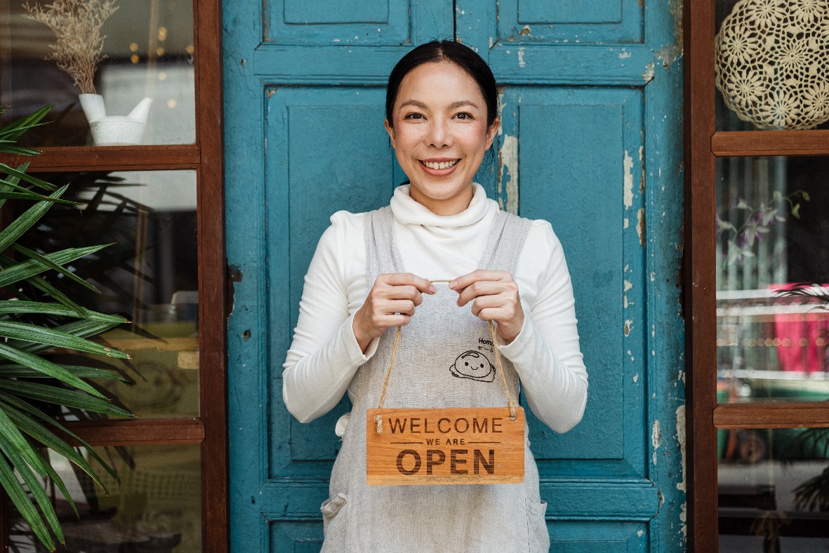 Photo of a woman standing in front of the door to her business holding a sign that says welcome we are open